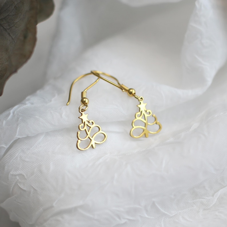 May include: A pair of gold-colored Christmas tree earrings. Each earring features a delicate, openwork design of a stylized tree with a star at the top. The earrings hang from simple, curved ear wires, set against a white, textured background.