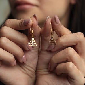 May include: Gold-colored Christmas tree earrings. Each earring features a stylized tree design with a star at the top and a looped base. The earrings are held up by a person's hands, showcasing the delicate details.