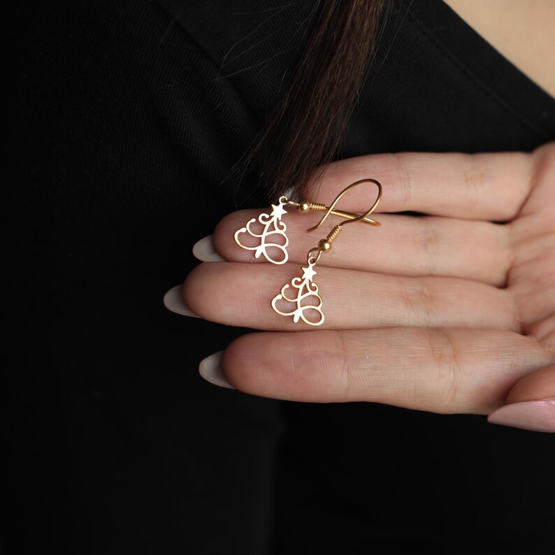 May include: A pair of gold-colored dangle earrings. Each earring features a stylized tree design with a star accent. The earrings are held in a hand against a black background.