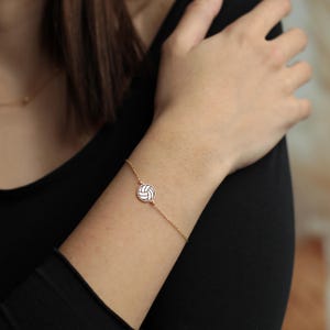 Peut inclure: Un bracelet dor&eacute; avec un pendentif rond repr&eacute;sentant un ballon de volley-ball blanc et noir. Le bracelet est port&eacute; au poignet, mettant en valeur un th&egrave;me sportif. La cha&icirc;ne est d&eacute;licate.