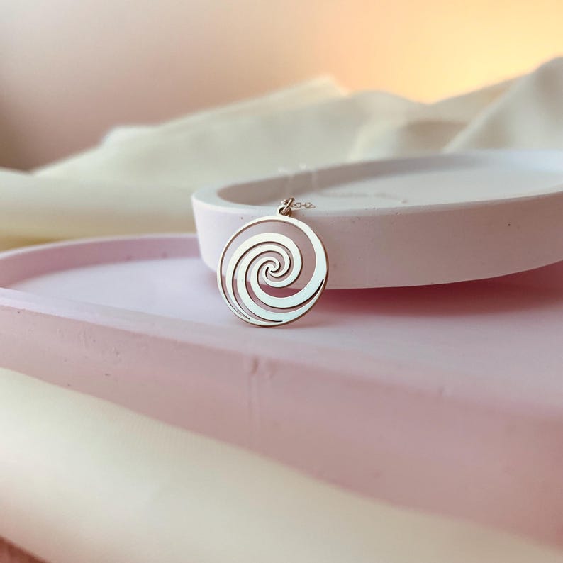 Puede incluir: Un collar de plata con un colgante circular que presenta un dise&ntilde;o en espiral. El colgante est&aacute; sobre una superficie blanca.