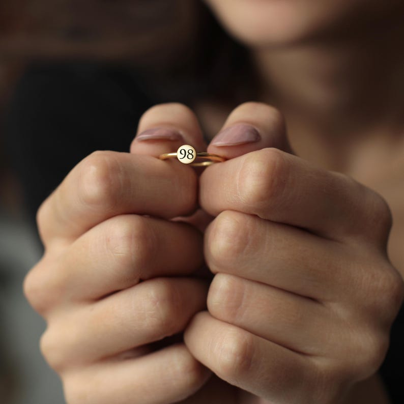 May include: A gold ring with a small, round, white face featuring the number 98 in black. The ring is held between two hands with manicured nails.