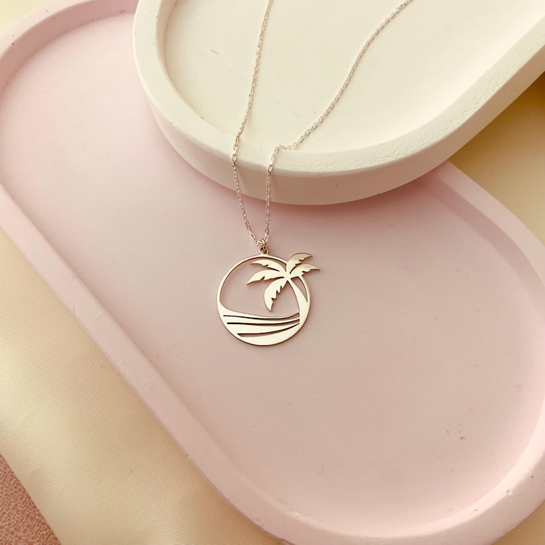 Puede incluir: Un collar de plata con un colgante de palmera y ola de mar. El colgante es un c&iacute;rculo con un dise&ntilde;o recortado de una palmera y olas del mar.