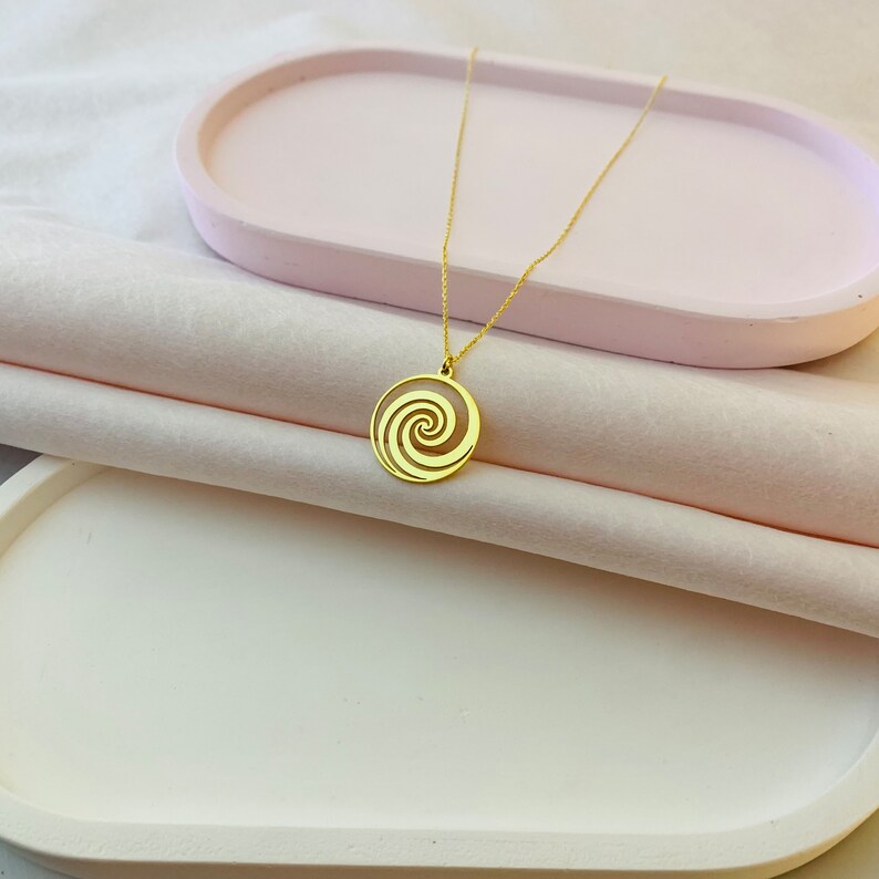 Puede incluir: Un collar de oro con un colgante circular que presenta un dise&ntilde;o en espiral. El colgante est&aacute; sobre una superficie blanca.