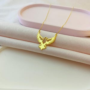 May include: A gold chain necklace with a pendant shaped like an eagle with its wings spread. The eagle is a cutout design, and the necklace is on a pink surface.