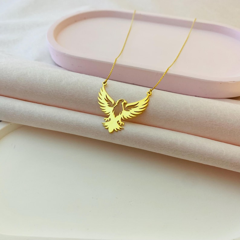 May include: A gold chain necklace with a pendant shaped like an eagle with its wings spread. The eagle is a cutout design, and the necklace is on a pink surface.
