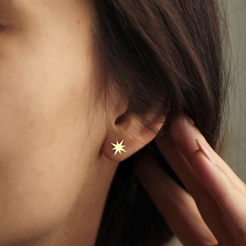K&ouml;nnte beinhalten: Nahaufnahme eines Ohrs mit einem goldfarbenen Stern-Ohrstecker. Der Ohrring ist klein und hat acht Zacken. Die Haut der Person ist hell, und ihr Haar ist dunkelbraun.