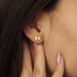May include: A close-up of a person's ear with a small, gold-colored bow-shaped earring. The earring is a stud style, and the bow design is detailed. The person's skin tone is fair, and their dark hair is visible.