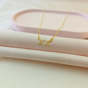 May include: A gold necklace featuring a triple moon pendant. The pendant is a delicate, open design with three crescent moons.