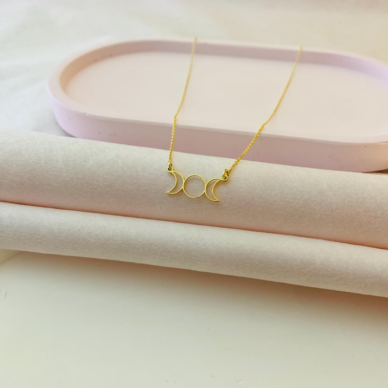 May include: A gold necklace featuring a triple moon pendant. The pendant is a delicate, open design with three crescent moons.