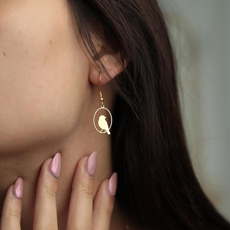 Puede incluir: Pendientes dorados con forma de p&aacute;jaro. Cada pendiente presenta una peque&ntilde;a silueta de p&aacute;jaro dentro de un marco circular, suspendida de un delicado gancho. Los pendientes se muestran en la oreja de una persona, destacando la elegante simplicidad del dise&ntilde;o y el tono c&aacute;lido del metal.