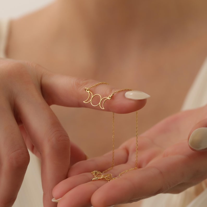 May include: A gold necklace featuring a triple moon pendant. The pendant is a simple design with three crescent moons.