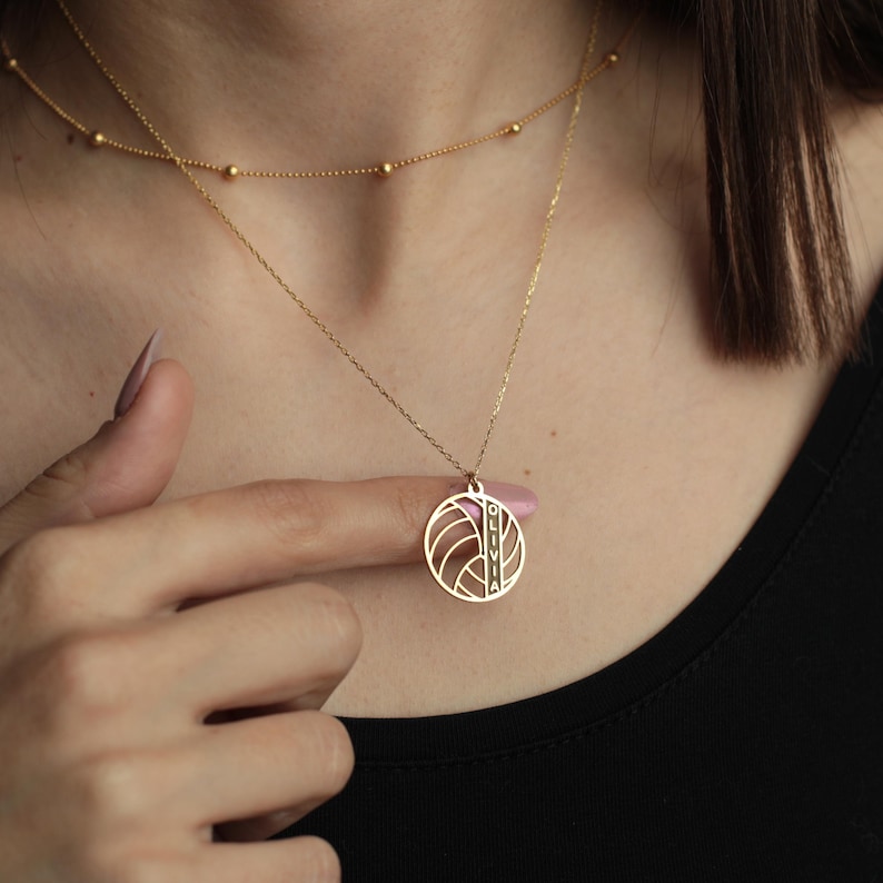 K&ouml;nnte beinhalten: Eine zarte goldene Halskette mit einem runden Anh&auml;nger mit geometrischem Design und dem eingravierten Namen "OLIVIA". Die Halskette ist mit einer zweiten, k&uuml;rzeren Kette mit kleinen goldenen Perlen versehen. Der Schmuck wird vor schwarzem Hintergrund pr&auml;sentiert.