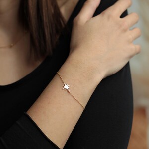 May include: A delicate gold-colored bracelet featuring a starburst charm. The bracelet is worn on a wrist, with the starburst charm positioned on the side. The bracelet is simple and elegant, suitable for everyday wear.