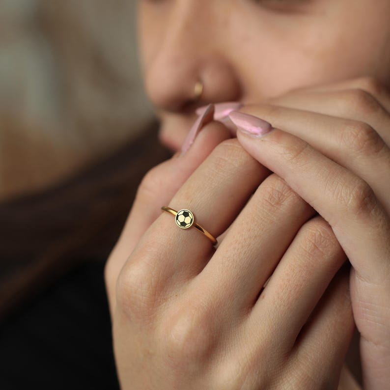 May include: A gold ring with a soccer ball design. The ring has a thin band and a small, circular charm with a black and white soccer ball pattern. The ring is worn on a finger.
