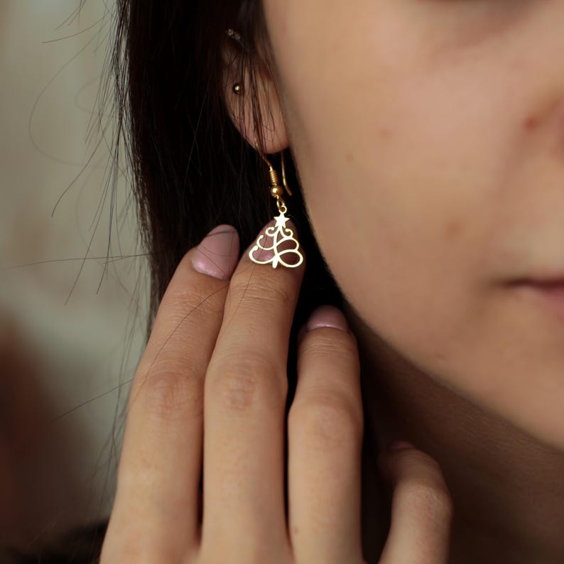 May include: A gold-colored Christmas tree earring with a star on top. The earring is a delicate, openwork design, hanging from a simple hook. The earring is shown close up, highlighting the intricate details of the festive jewelry.