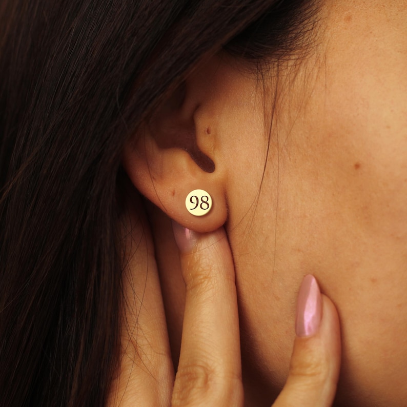 May include: A gold-coloured stud earring featuring the number "98" in a circular design. The earring is worn on an ear. The image shows a close-up of the ear and part of the face.