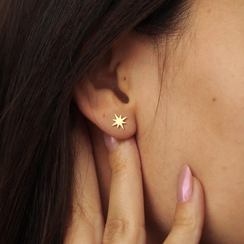 K&ouml;nnte beinhalten: Nahaufnahme eines Ohrs mit einem kleinen, goldfarbenen Stern-Ohrstecker. Der Ohrring hat acht Zacken und ist auf der Haut angebracht. Im Hintergrund sind die dunklen Haare der Person sowie eine Hand zu sehen, die das Ohr ber&uuml;hrt.