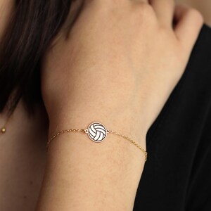 Peut inclure: Un bracelet dor&eacute; orn&eacute; d'un charm circulaire repr&eacute;sentant un ballon de volley-ball. Le ballon est blanc avec des lignes noires. Le bracelet est port&eacute; sur un poignet, avec un haut noir en arri&egrave;re-plan.