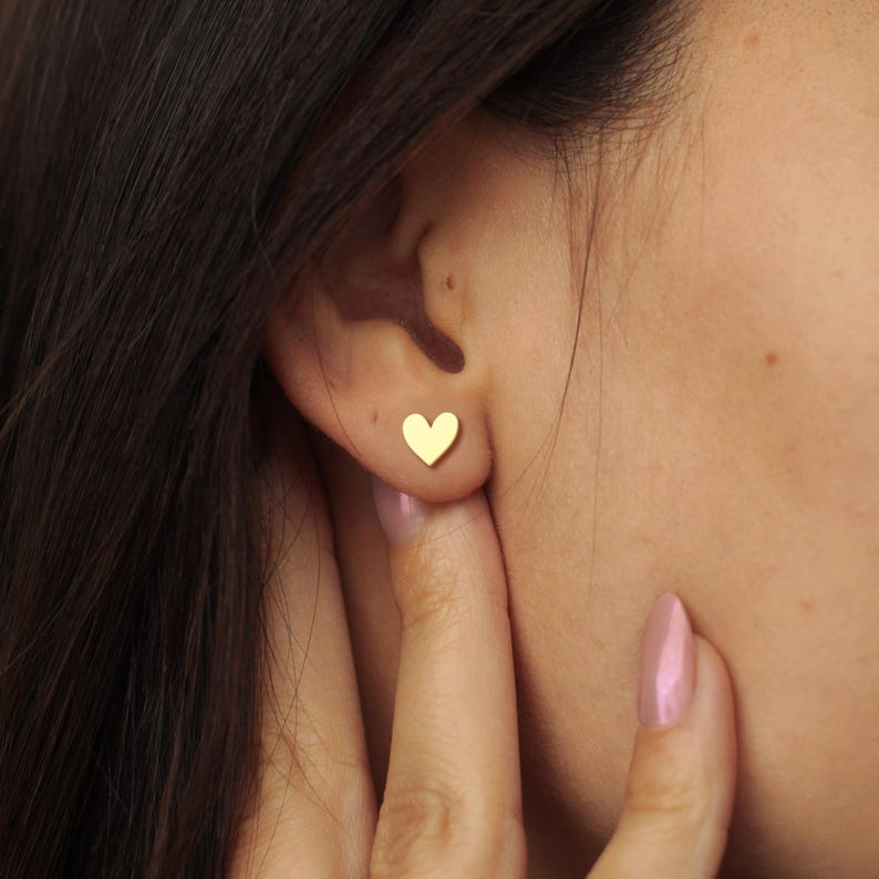May include: A close-up of a person's ear with a small, gold heart-shaped stud earring. The earring is a simple, minimalist design. The person's skin is fair, and their dark hair is visible in the background.