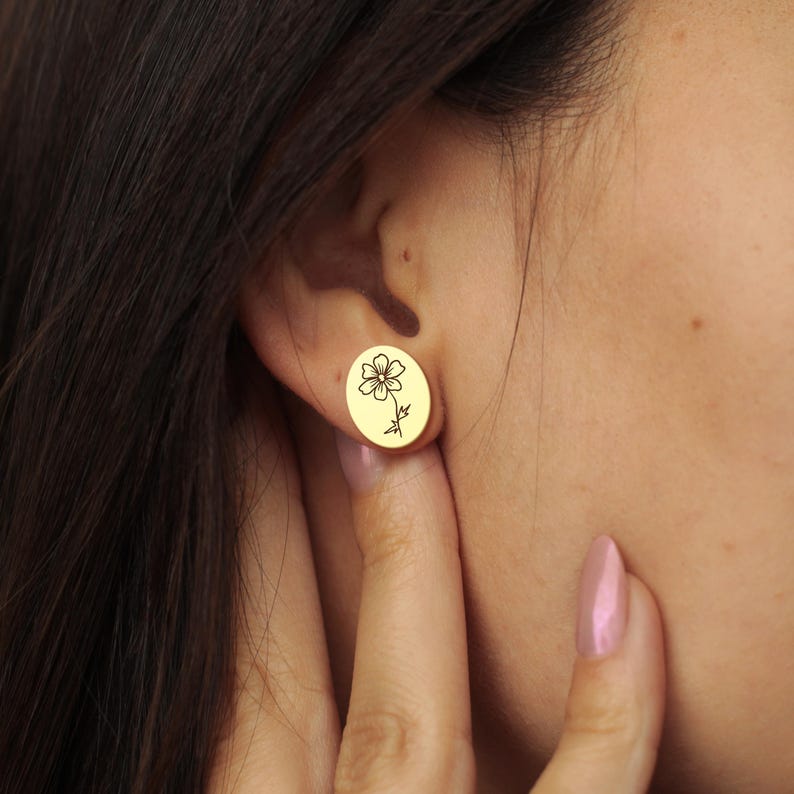 May include: Gold-colored oval stud earrings. Each earring features a delicate black floral design. The earrings are being worn, showcasing their size and style. A simple and elegant accessory.
