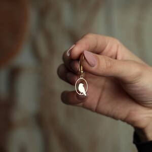 Puede incluir: Pendiente dorado con la silueta de un p&aacute;jaro dentro de un marco ovalado. El pendiente tiene un dise&ntilde;o de gancho simple. El p&aacute;jaro est&aacute; representado en un estilo minimalista, con un cuerpo curvo y una peque&ntilde;a cola.