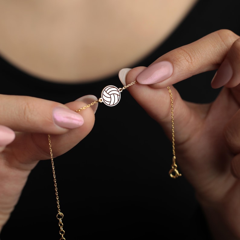 Peut inclure: Un bracelet dor&eacute; avec un charm en forme de ballon de volley-ball. Le charm est un pendentif circulaire avec un motif d&eacute;taill&eacute; de ballon de volley-ball. Le bracelet est tenu entre deux mains avec du vernis &agrave; ongles rose sur fond noir.