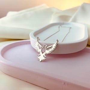 May include: A silver necklace with a delicate eagle pendant. The eagle is in flight with its wings spread wide. The necklace is on a white surface.