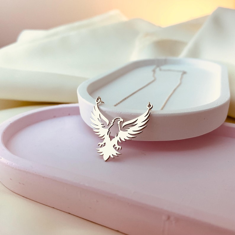 May include: A silver necklace with a delicate eagle pendant. The eagle is in flight with its wings spread wide. The necklace is on a white surface.