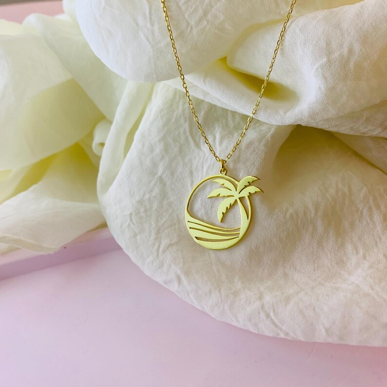 Puede incluir: Collar de oro con un colgante de palmera y escena de oc&eacute;ano. El colgante es un c&iacute;rculo con un dise&ntilde;o recortado de una palmera y olas.