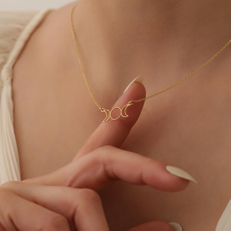 May include: A gold necklace with a delicate crescent moon pendant. The pendant features three moons in different phases, symbolising the cycle of the moon.