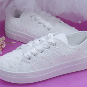 Può includere: Sneakers bianche ricoperte di pizzo con suola platform. Le scarpe presentano lacci in nastro di raso e sono decorate con piccoli accenti di perle. Il design include un motivo floreale in pizzo. Sfondo rosa.