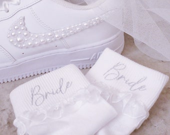 Könnte beinhalten: Weiße Brautsocken mit Spitzenbesatz und dem Wort "Bride" in silbernen Glitzer gestickt. Die Socken werden mit weißen Sneakers kombiniert, die mit Perlen verziert sind.