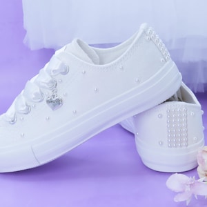 Puede incluir: Zapatillas de lona blancas adornadas con perlas y un dije en forma de corazón que dice "Flower Girl". Los zapatos tienen cordones de cinta de raso y detalles de perlas en la puntera y el talón. Sobre un fondo morado suave.