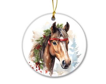 May include: A round white ceramic ornament featuring a brown horse with a white blaze on its face. The horse is adorned with a red ribbon headband and a garland of green pine branches with red berries. A gold string is attached for hanging.
