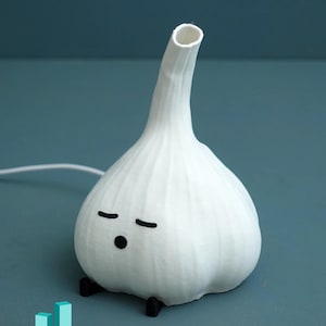 May include: A whimsical white garlic-shaped lamp with a long neck and a small opening at the top. The lamp has a cartoon face with closed eyes and a small black dot for a mouth. It sits on two small black feet.