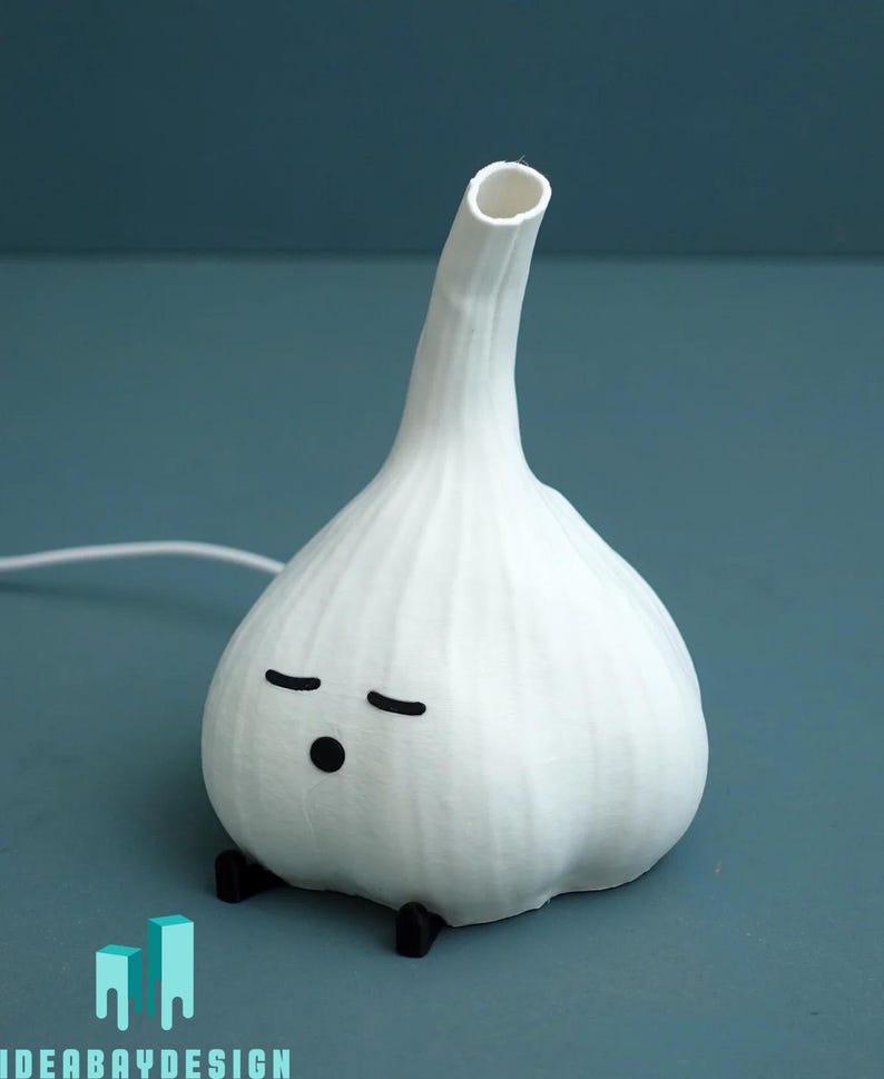 May include: A whimsical white garlic-shaped lamp with a long neck and a small opening at the top. The lamp has a cartoon face with closed eyes and a small black dot for a mouth. It sits on two small black feet.