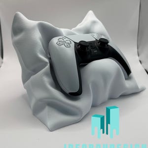 Pode incluir: Um controlador de jogos branco descansando sobre um travesseiro branco. O controlador tem detalhes e botões pretos. O travesseiro é feito de um tecido macio e felpudo. A imagem é rotulada com "IDEABAYDESIGN".