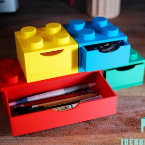 Könnte beinhalten: Ein Satz von vier bunten, stapelbaren Schubladen in Form von Lego-Steinen. Die Schubladen sind rot, gelb, blau und grün. Die oberste Schublade ist offen und zeigt einen kleinen Gegenstand im Inneren.