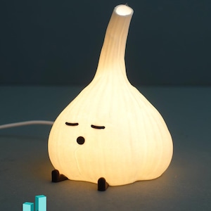 Op de afbeelding: Een grillige, oplichtende knoflookvormige lamp met een cartoongezicht, zwarte voetjes en een steel. De lamp straalt een zacht, warm licht uit en creëert een gezellige sfeer. De tekst "IDEABAYDESIGN" is zichtbaar.