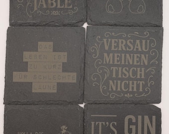 May include: Set of six black slate coasters with various humorous sayings and designs. The coasters feature sayings like "Don't Fuck Up The Table", "Everybunny Needs Somebunny", "Das Leben Ist Zu Kurz Für Schlechte Laune", "Versau Meinen Tisch Nicht", "Holla Die Weinfee", and "It's Gin O'Clock".