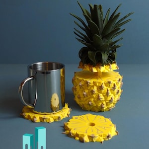 Könnte beinhalten: Satz mit vier gelben Untersetzern in Ananasform, auf einem der Untersetzer steht eine Metalltasse. Die Ananas-Untersetzer haben eine strukturierte Oberfläche und ein Loch in der Mitte. Die Tasse ist silberfarben und hat einen Griff.