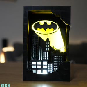 May include: A black Batman-themed night light depicting a cityscape, illuminated from within. The design includes a yellow Bat-Signal and a detailed city silhouette. The light is housed in a rectangular frame with a distressed brick effect. The text "IDEABAYDESIGN" is visible.