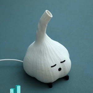 May include: A whimsical white garlic-shaped lamp with a curved stem and a cartoon face with closed eyes. The lamp has small black feet and a white cord. The design is a playful take on a kitchen staple.