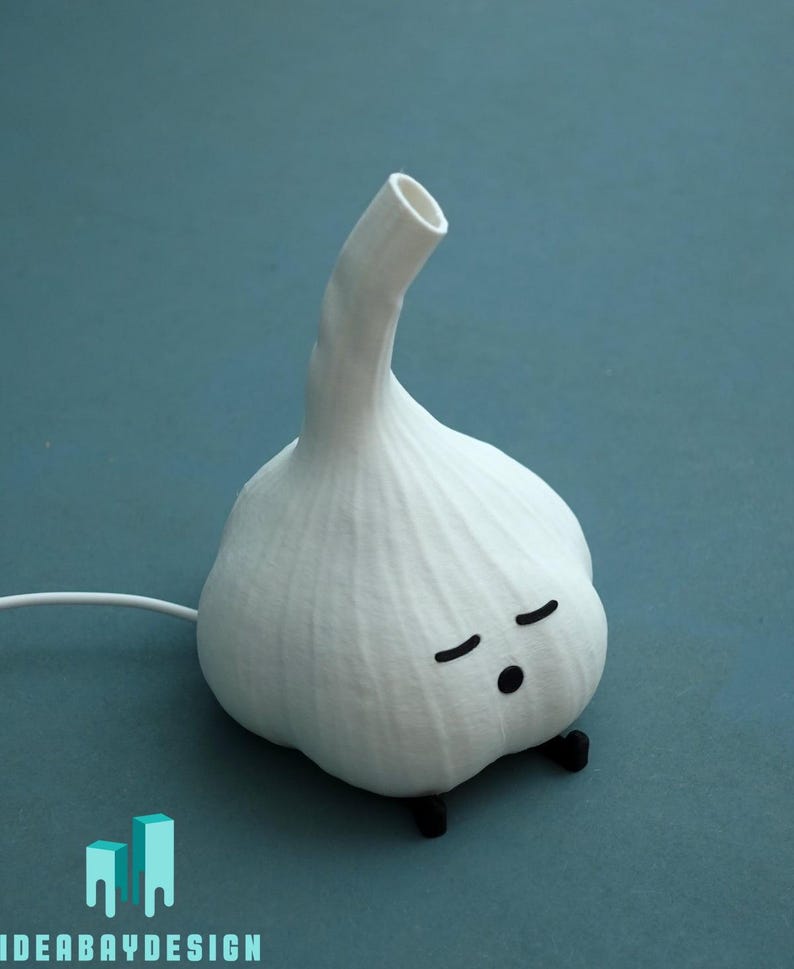 May include: A whimsical white garlic-shaped lamp with a curved stem and a cartoon face with closed eyes. The lamp has small black feet and a white cord. The design is a playful take on a kitchen staple.