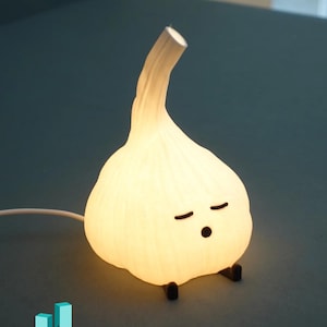 May include: A whimsical, glowing garlic-shaped lamp with a cartoon face, featuring closed eyes and a small mouth. The lamp emits a soft, warm light and has small black feet. The design is playful and unique, perfect for a bedroom or desk.