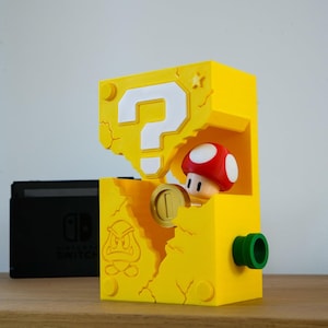 Op de afbeelding: Een gele, 3D-geprinte vraagtekenbox met een rood-witte paddenstoelfiguur en een gouden munt. De doos heeft een groene cilinder aan de zijkant en een Bowser-afbeelding. Een zwarte Nintendo Switch-console is zichtbaar op de achtergrond.