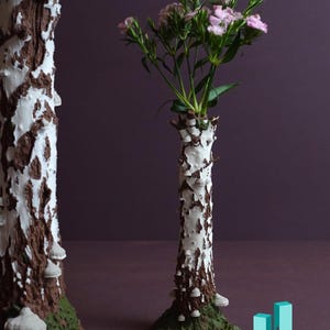 May include: Two white and brown ceramic vases shaped like birch trees. The smaller vase has pink flowers in it. The vases are on a green surface against a purple background.