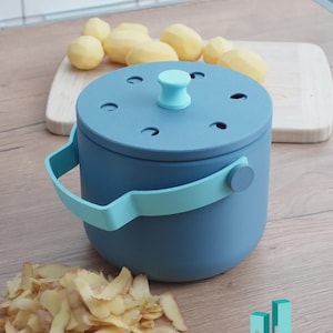May include: A blue and teal compost bin with a handle and lid, next to a pile of potato peelings. The lid has several small holes and a teal handle. Peeled potatoes and a wooden board are in the background.