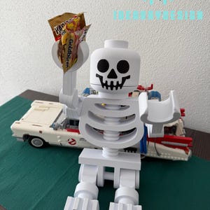 May include: A white plastic skeleton figure holding a bag of sweets. The skeleton is wearing a black skull face and is sitting down with its legs crossed. The figure is holding the sweets in its right hand.